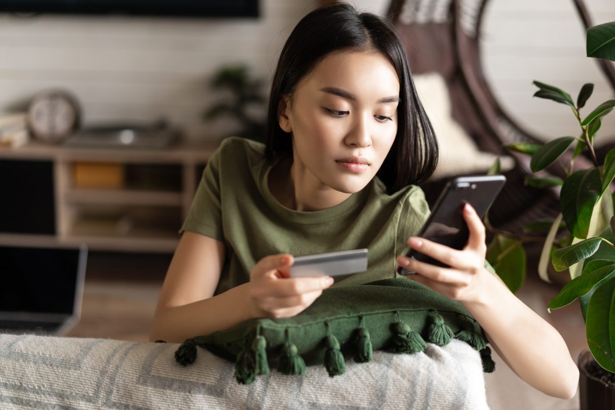 Online shopping. Asian woman purchase online, shopping in application on mobile phone, using credit card info to pay, sitting at home in living room.